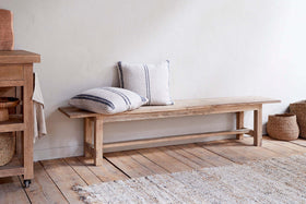 The nkuku Fioni Reclaimed Wood Bench - Natural sits against a white wall on wooden flooring, paired with two striped, neutral pillows. Woven baskets and a textured rug complete the cozy, minimalist farmhouse look.