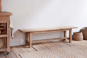 The Fioni Reclaimed Wood Bench - Natural by nkuku sits against a white wall on a rustic floor, flanked by woven baskets and a wooden cabinet, while a textured beige zigzag rug enhances the farmhouse look.