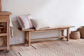 The nkuku Fioni Reclaimed Wood Bench - Natural sits against a white wall on a wooden floor, topped with two textured throw pillows. Woven baskets and a beige geometric rug complete the farmhouse look in front of the bench.