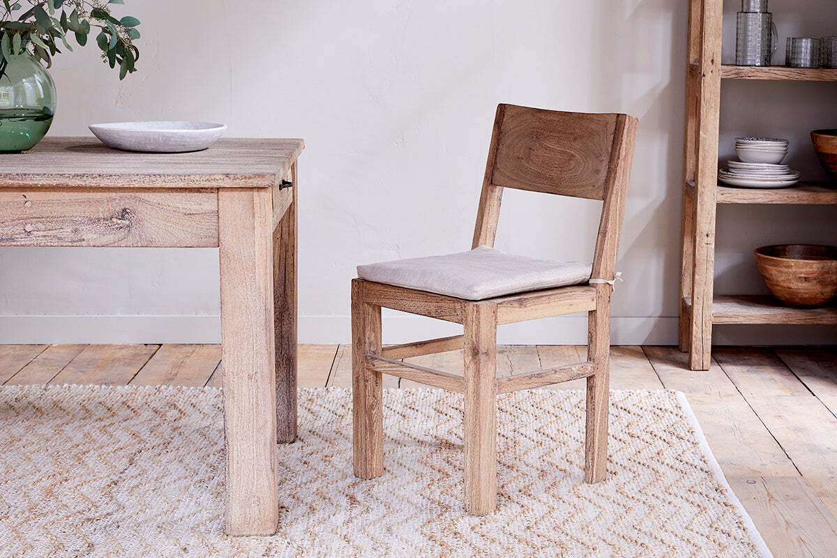 The Fioni Reclaimed Wood Dining Chair - Natural by nkuku, featuring a beige cushion, sits beside a matching table on a light rug. Farmhouse touches like shelves with bowls and plates plus a green vase create a cozy dining atmosphere.