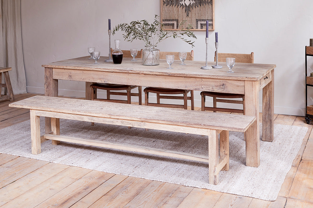 The nkuku Fioni Reclaimed Wood Dining Table – Natural is paired with a matching bench and chairs on a light rug, set with glassware, a carafe, candlesticks, and a vase of greenery. Wooden floors and neutral walls create a cozy, minimalist space.