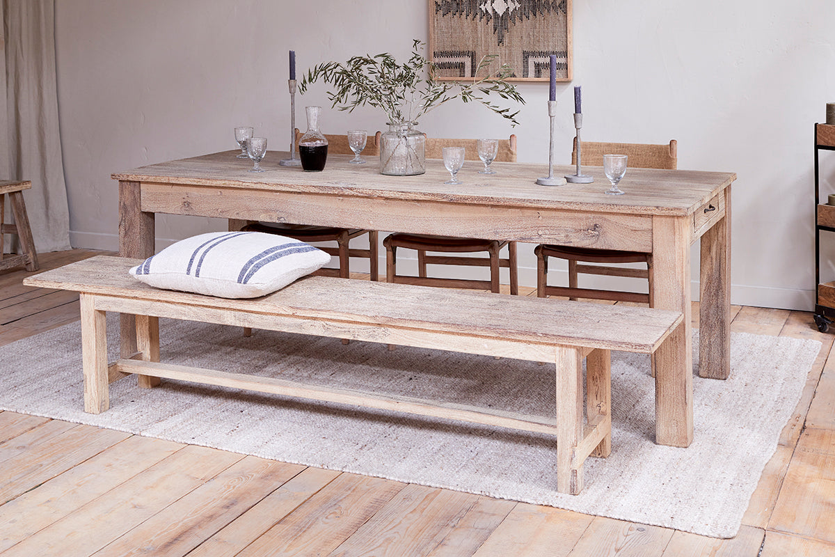The nkuku Fioni Reclaimed Wood Dining Table – Natural is styled with four chairs, a matching bench with a striped pillow, set with glasses, a carafe, candles, and olive branches. Light wood floors, a white rug, and neutral decor complete the look.