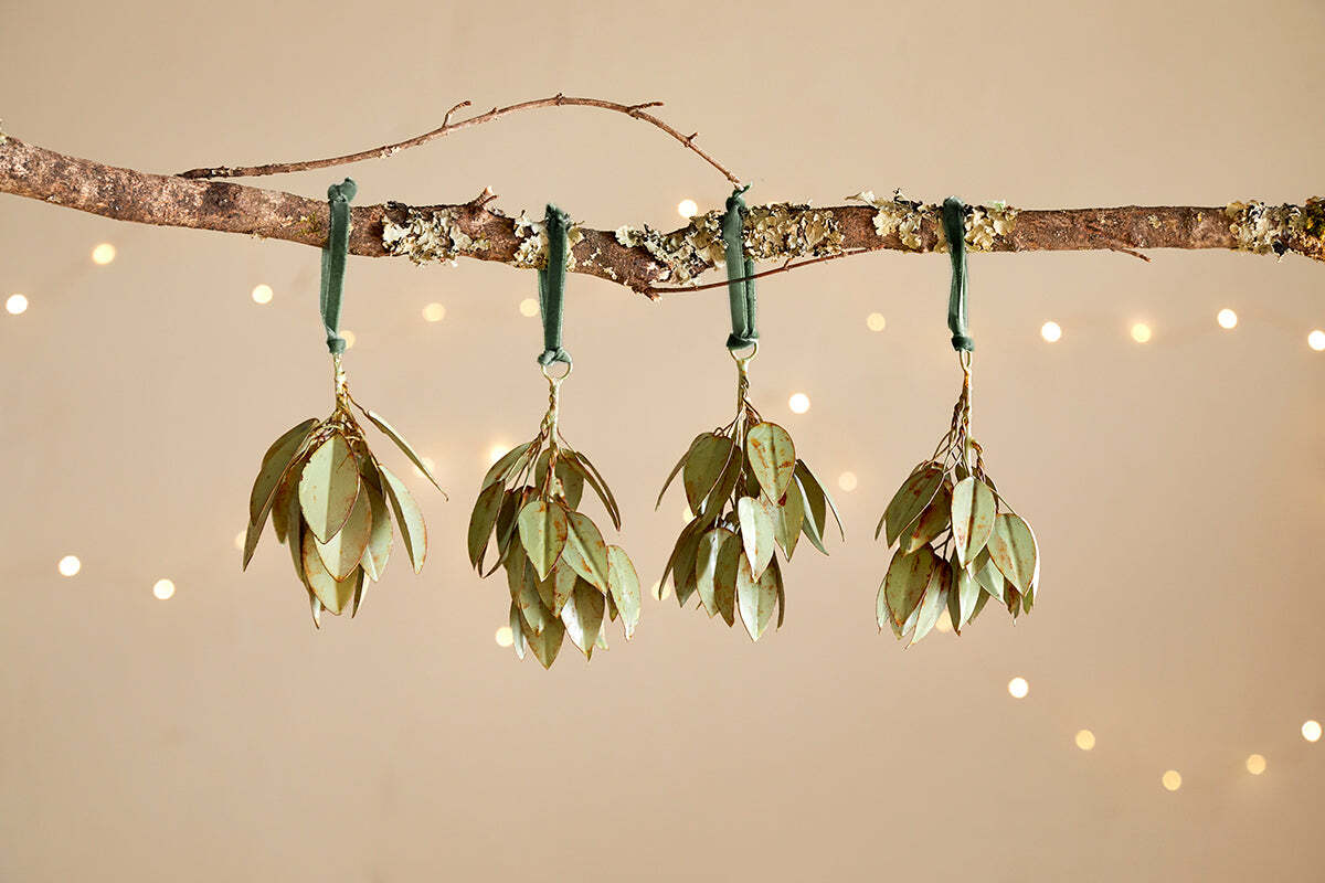 Four Geeti Baubles - Soft Green by nkuku, with gold-edged green leaves, hang from a branch on green ribbons. Set against a beige backdrop with soft string lights, they bring a warm, festive glow to your decor.