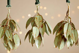 Four Geeti Baubles in soft green by nkuku hang from green velvet ribbons against a beige backdrop with blurred golden lights, adding a nature-inspired, serene touch with their elegant curves and subtle, organic hue.