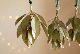 Four Geeti Baubles - Soft Green by nkuku hang from green ribbons against a soft beige background, with warm, blurred fairy lights. Their pale green finish and earthy tones evoke nature-inspired ornaments in this tranquil setting.