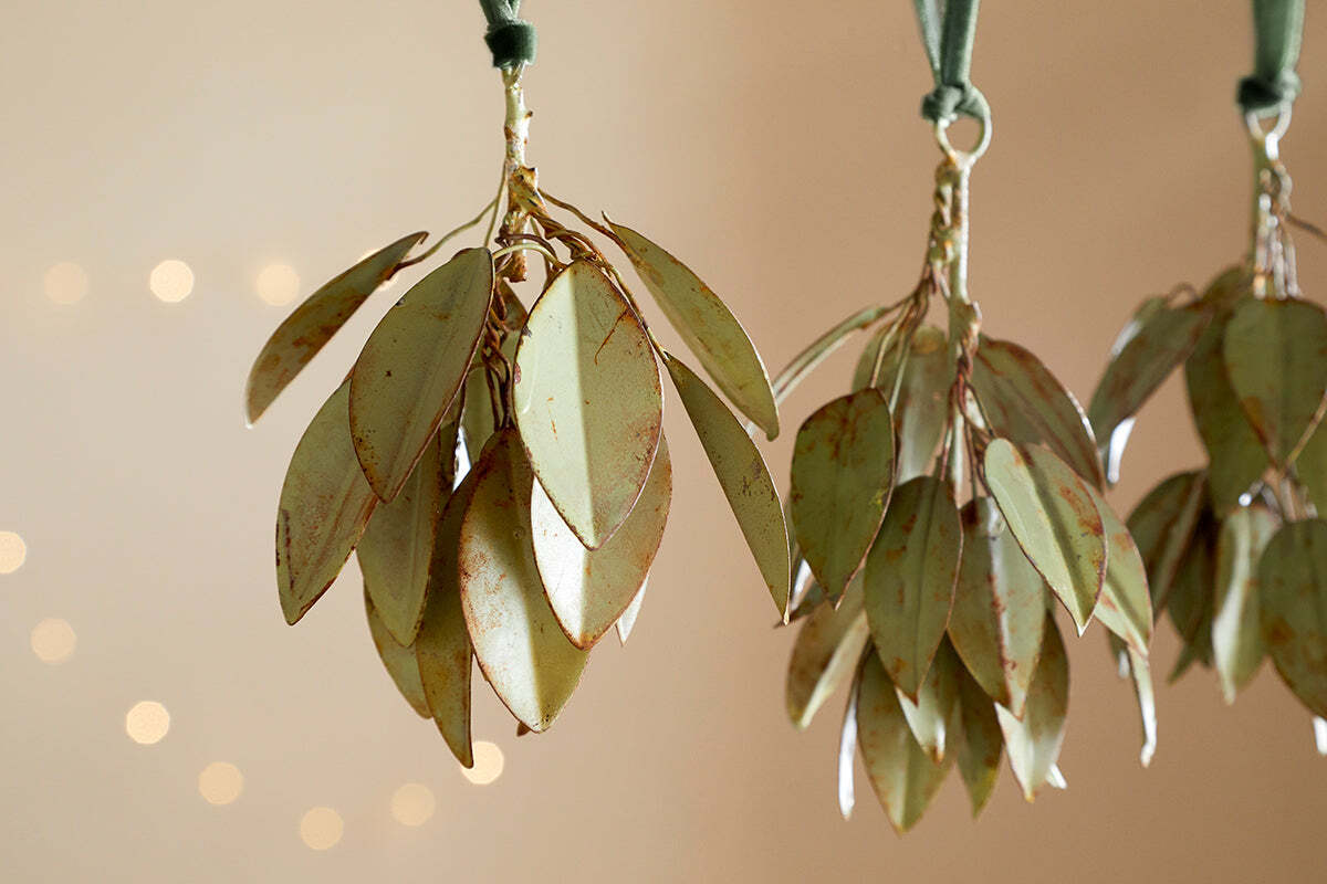 Four Geeti Baubles - Soft Green by nkuku hang from green ribbons against a soft beige background, with warm, blurred fairy lights. Their pale green finish and earthy tones evoke nature-inspired ornaments in this tranquil setting.