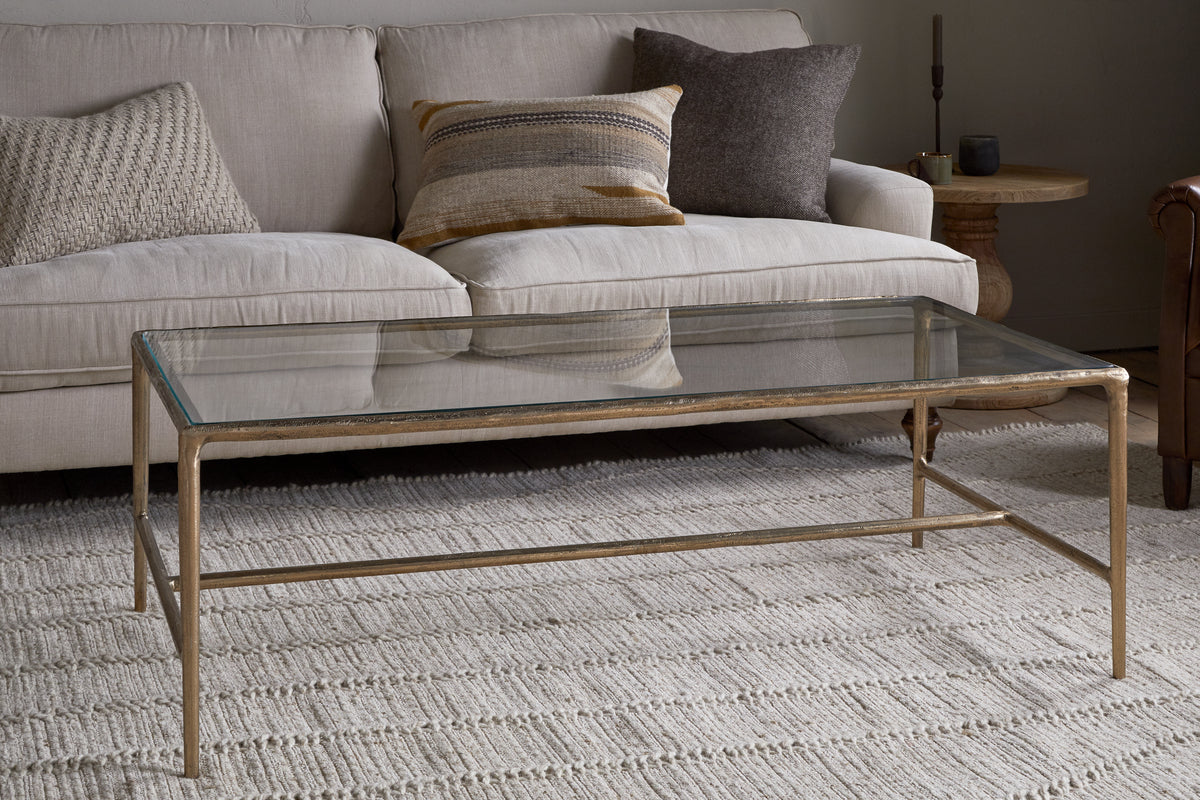 The nkuku Gilasi Cast Metal & Glass Coffee Table in antique brass sits on a textured cream rug before a light beige sofa with patterned cushions, creating a cozy living room setting.