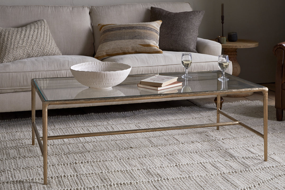The nkuku Gilasi Cast Metal & Glass Coffee Table in Antique Brass displays a white textured bowl, two glasses of white wine, and a book. Behind it is a beige sofa with neutral cushions on a textured rug.