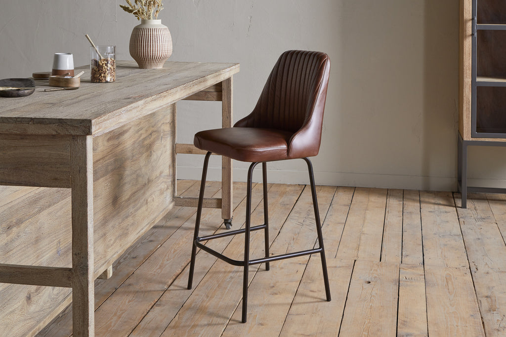 The nkuku Harsha Leather Counter Chair in chocolate brown, featuring black metal legs, complements neutral decor and rustic wooden tables—ideal for minimalist kitchen island seating on a light wood floor.