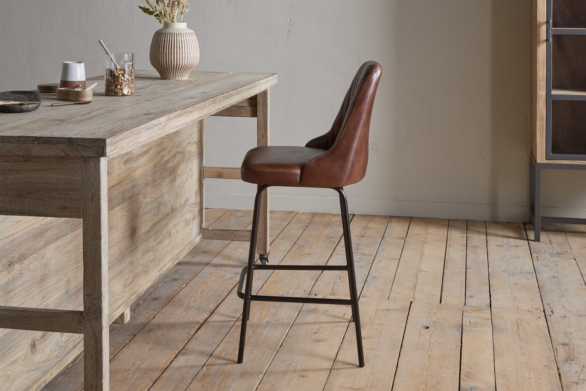 The nkuku Harsha Leather Counter Chair in chocolate brown, featuring black metal legs, provides stylish seating beside a rustic wooden table on a light wood floor in a minimalist room with neutral walls.