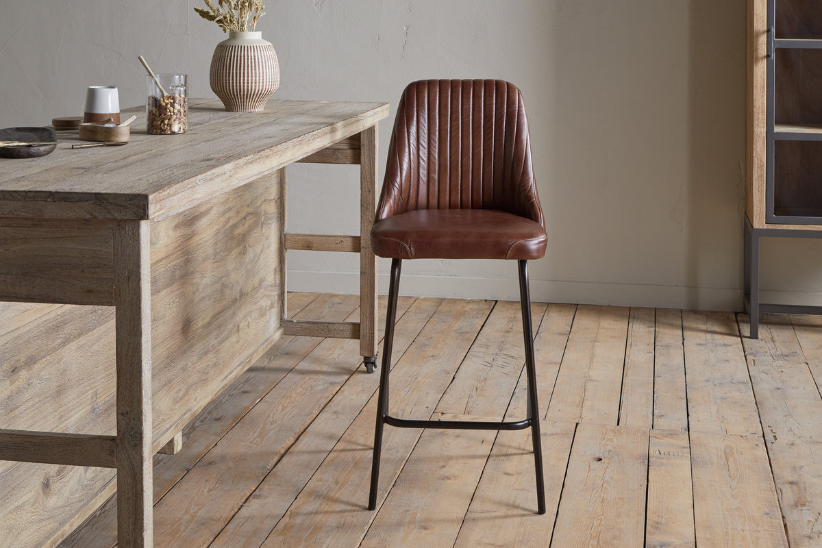 A modern kitchen features a wooden table on the left, ceramic vases and bowls, and the nkuku Harsha Leather Counter Chair in chocolate brown with black metal legs offering stylish seating at the island on a light wooden floor.
