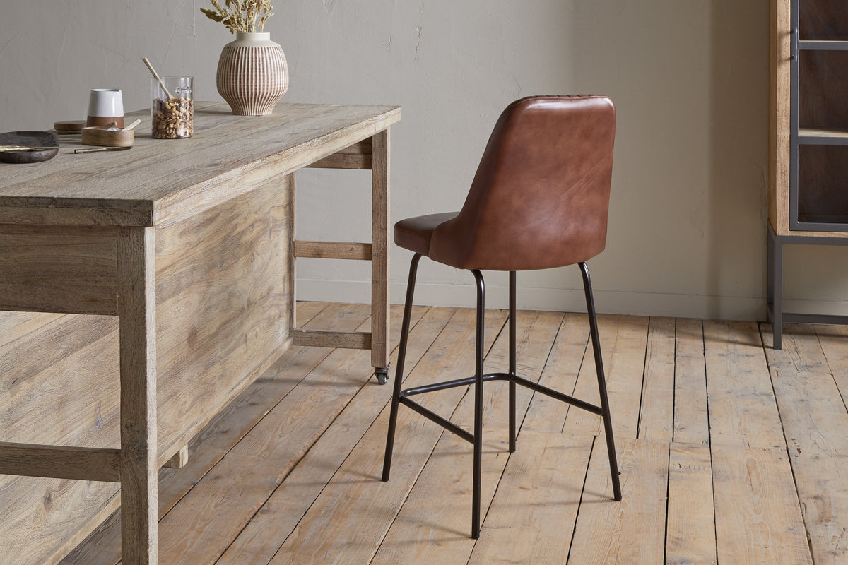 The Harsha Leather Counter Chair in chocolate brown by nkuku, featuring black metal legs, sits beside a rustic wooden table on light wood floors with neutral walls—ideal for stylish kitchen island seating. The table is adorned with a vase of dried flowers and jars.