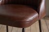 Close-up of the Harsha Leather Counter Chair by nkuku in chocolate brown, featuring visible stitching, curved edges, and black metal legs—ideal for kitchen island seating and shown on a wooden floor.