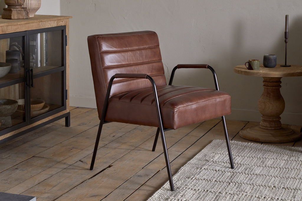 The Hadithi Leather Occasional Chair in chocolate brown by nkuku, featuring black metal legs, is placed on a wooden floor beside a woven rug. A glass-door wooden cabinet and round side table with candle and cup enhance the cozy space.