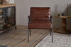The Hadithi Leather Occasional Chair by nkuku, in chocolate brown buffalo leather with black metal legs, rests on a wooden floor near a patterned rug, next to a wooden side table and a glass-doored cabinet with pottery inside.