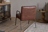 The nkuku Hadithi Leather Occasional Chair in chocolate brown, featuring a ribbed design and metal legs, sits on a light wooden floor near a beige textured rug alongside a wooden side table and glass-front cabinet against neutral walls.