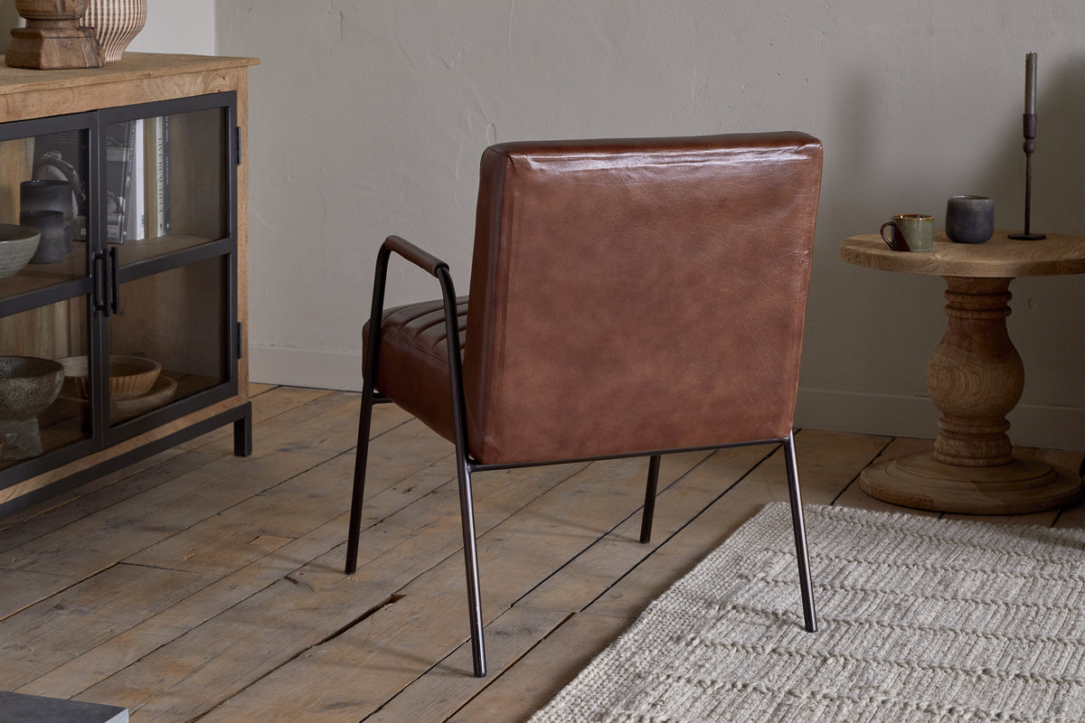 The nkuku Hadithi Leather Occasional Chair in chocolate brown, featuring a ribbed design and metal legs, sits on a light wooden floor near a beige textured rug alongside a wooden side table and glass-front cabinet against neutral walls.