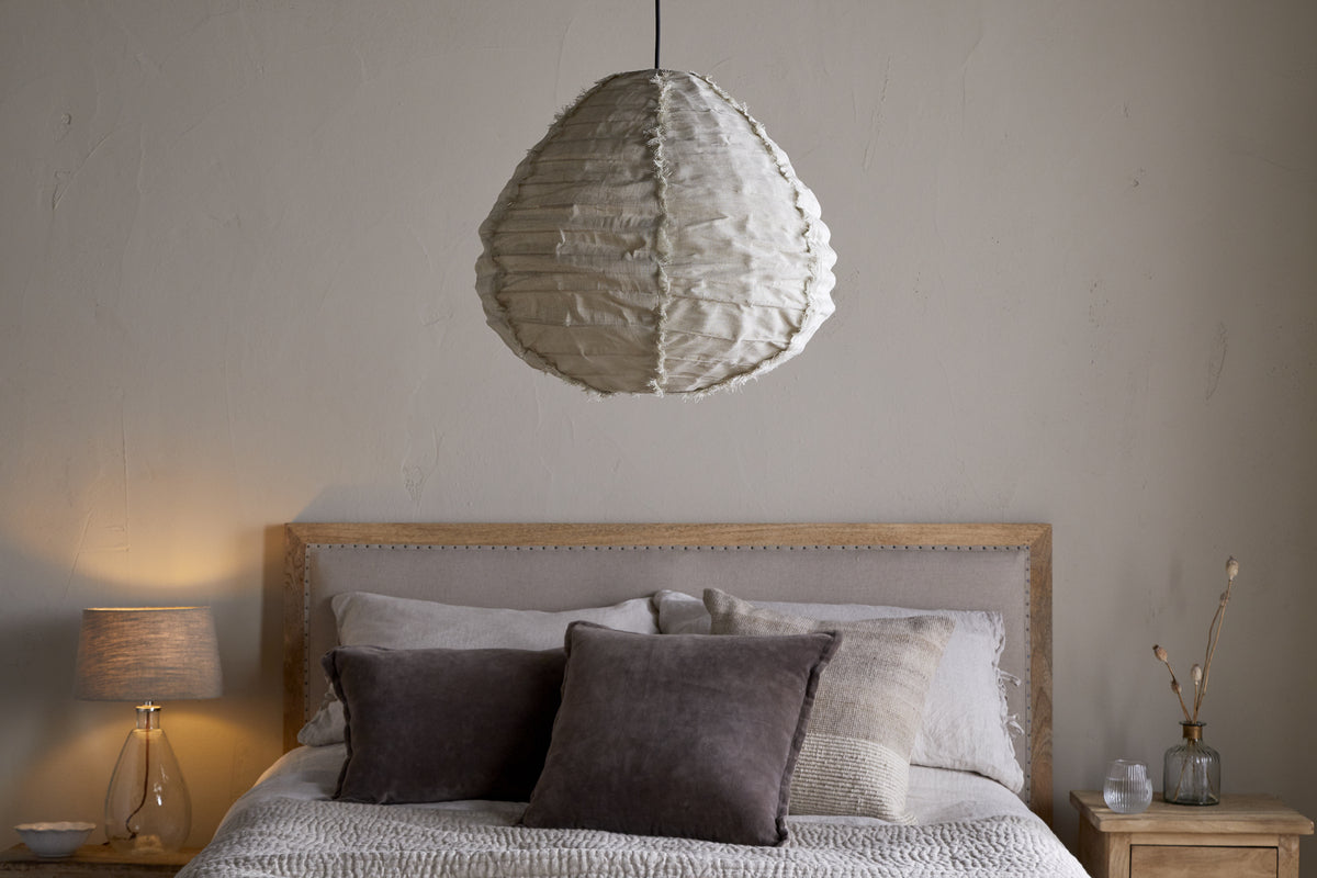 A cozy bedroom features a light wood bed frame, neutral bedding with two dark pillows, and the nkuku Hirana Linen Lampshade (Natural, Small) overhead. A wooden nightstand holds a lamp, vase, and minimal decor against a beige wall.