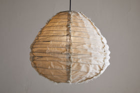 The Hirana Linen Lampshade by nkuku in natural, small size hangs from the ceiling, its textured shade softly glowing against a plain light gray background.