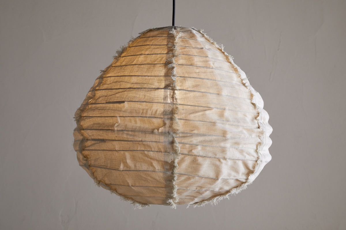 The Hirana Linen Lampshade by nkuku in natural, small size hangs from the ceiling, its textured shade softly glowing against a plain light gray background.