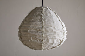The Hirana Linen Lampshade - Natural - Small by nkuku, shaped like a teardrop with textured off-white linen, visible seams, and frayed edges, hangs from a slim black cord—an ideal bedroom pendant light against a plain wall.