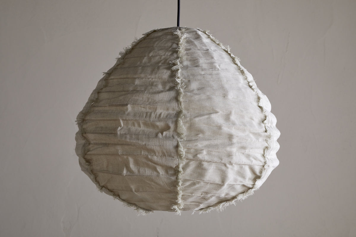 The Hirana Linen Lampshade - Natural - Small by nkuku, shaped like a teardrop with textured off-white linen, visible seams, and frayed edges, hangs from a slim black cord—an ideal bedroom pendant light against a plain wall.