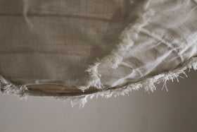 Close-up of the edge of the Hirana Linen Lampshade - Natural - Small by nkuku, showing its beige, textured linen fabric with visible fibers and loose threads against a soft, neutral background.