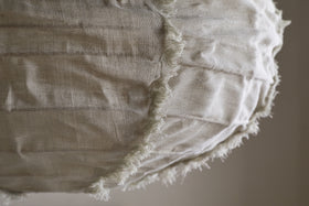 Close-up of the nkuku Hirana Linen Lampshade - Natural - Small, with visible seams and frayed edges. Its off-white textured linen and stitching create a rustic, handmade look—ideal for bedroom or pendant lighting. Background is softly blurred.