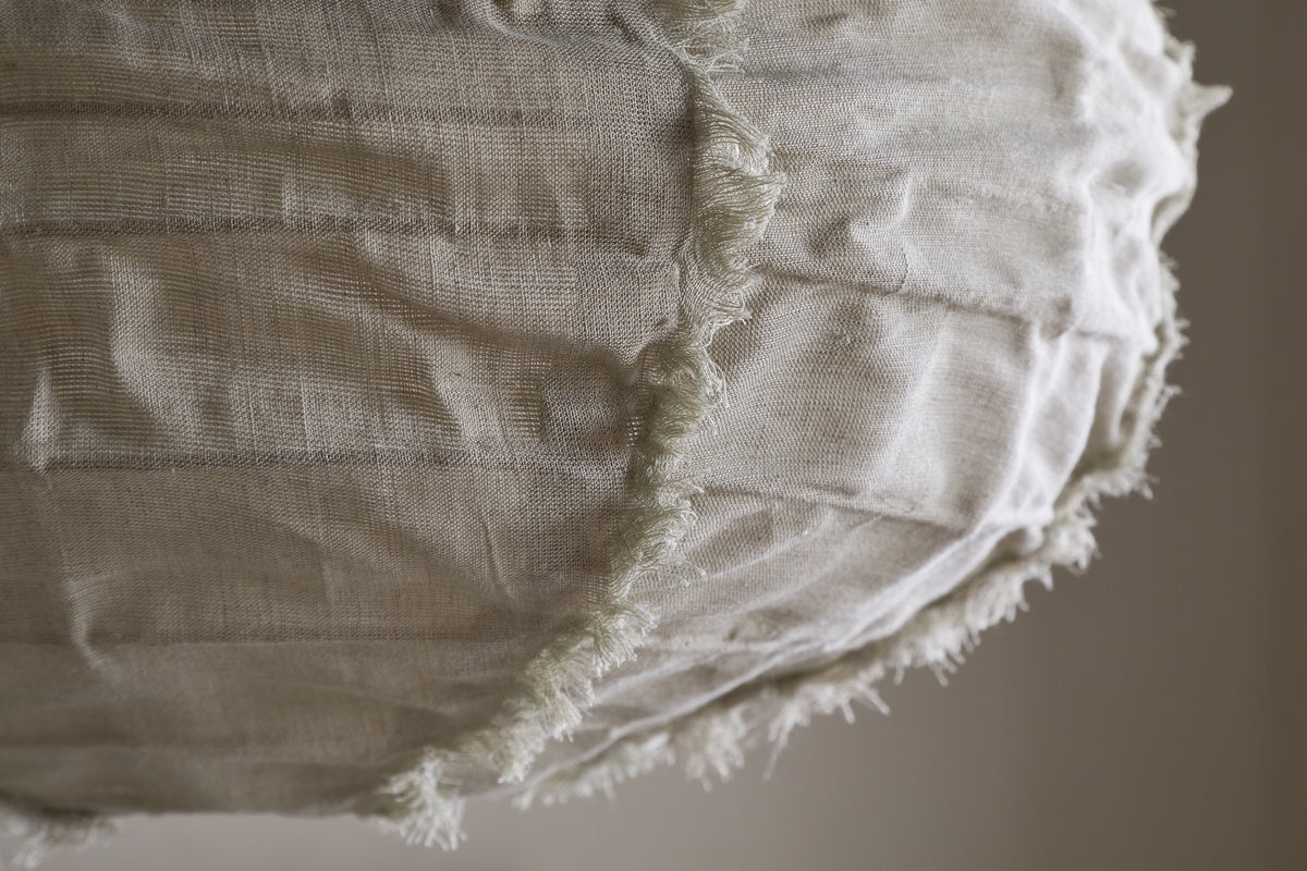 Close-up of the nkuku Hirana Linen Lampshade - Natural - Small, with visible seams and frayed edges. Its off-white textured linen and stitching create a rustic, handmade look—ideal for bedroom or pendant lighting. Background is softly blurred.