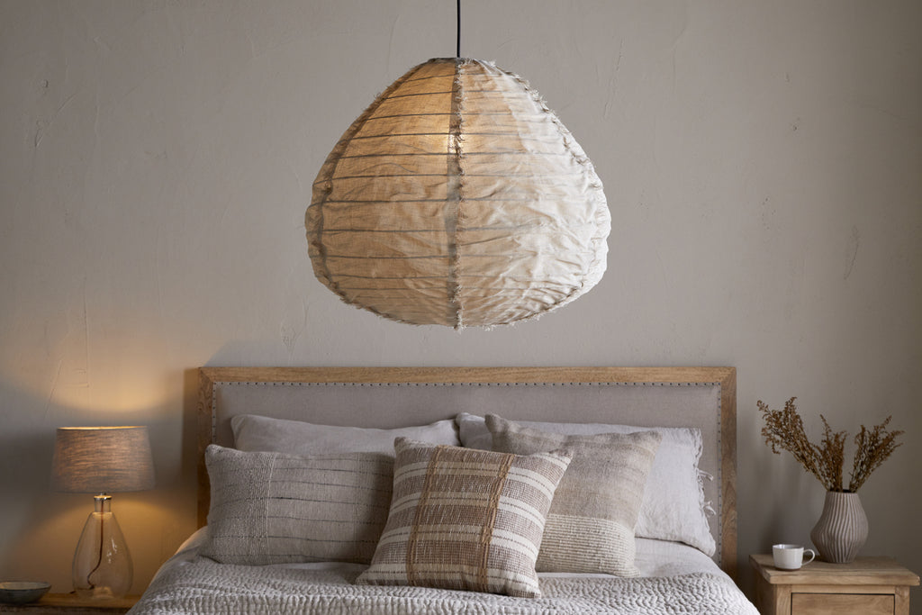 A cozy bedroom features the nkuku Hirana Cotton Lampshade - Natural - Large above the bed. Neutral pillows and bedding dress the bed, while a wooden nightstand with a lamp and dried flowers adds charm to the space.