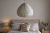 A cozy bedroom features the large Hirana Cotton Lampshade in natural by nkuku as a pendant above the bed, paired with neutral bedding, decorative pillows, and a wooden nightstand topped with a lamp and dried flowers.