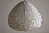 The nkuku Hirana Cotton Lampshade - Natural (Large) is a teardrop pendant with a textured beige cotton shade, visible seams, and frayed edges—ideal as a unique bedroom light against a plain wall.