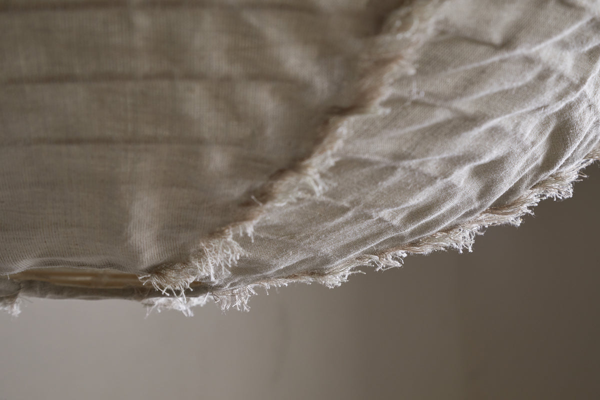 Close-up of the Hirana Cotton Lampshade by nkuku in natural, large size: off-white textured cotton with frayed edges and visible stitching, softly draped—perfect for adding warmth to any bedroom or living space.