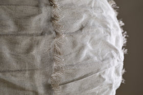 Close-up of the Hirana Cotton Lampshade by nkuku in Natural (Large), showing its light beige textured cotton fabric with a vertical seam and frayed edges, softly illuminated to highlight its unique details.