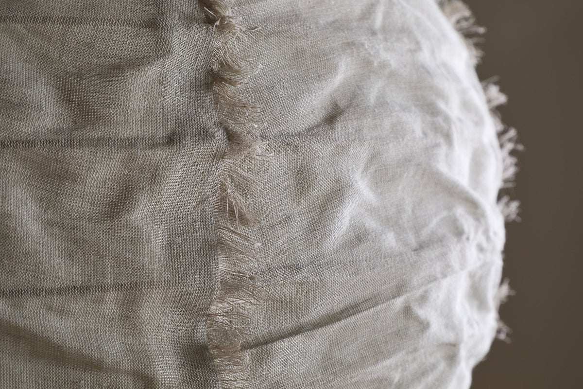 Close-up of the Hirana Cotton Lampshade by nkuku in Natural (Large), showing its light beige textured cotton fabric with a vertical seam and frayed edges, softly illuminated to highlight its unique details.