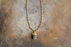 The nkuku Hadari Labradorite Necklace - Gold features a rough, irregular labradorite pendant set in gold, shown on an earthy brown textured stone surface.