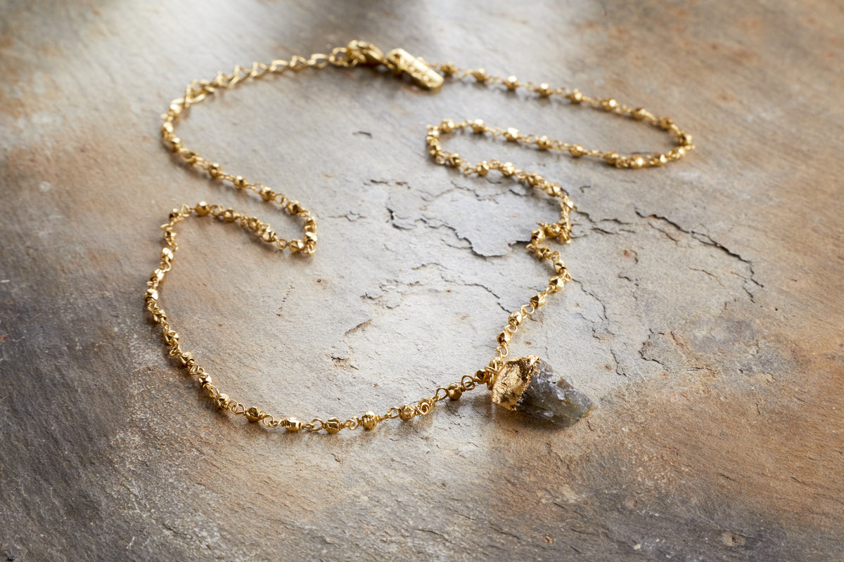 The nkuku Hadari Labradorite Necklace - Gold features a rough gray labradorite stone and delicate bead details, displayed on a textured rustic brown surface.