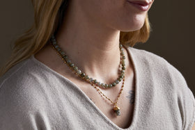 A woman in a beige top showcases two layered necklaces: the nkuku Hadari Labradorite Necklace - Gold, featuring round greenish stones, and a gold chain with a small pendant. A partial tattoo can be seen on her chest.