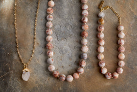 Three elegant jewellery pieces on a stone surface: nkuku’s Hadari Rose Quartz Necklace – Gold, with a gold chain and rose quartz pendant, alongside two beaded necklaces in pink and cream marbled stones with gold accents.