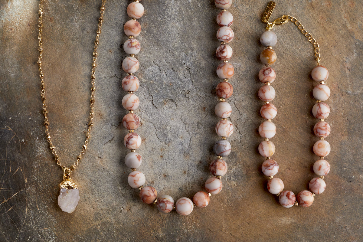 Three elegant jewellery pieces on a stone surface: nkuku’s Hadari Rose Quartz Necklace – Gold, with a gold chain and rose quartz pendant, alongside two beaded necklaces in pink and cream marbled stones with gold accents.