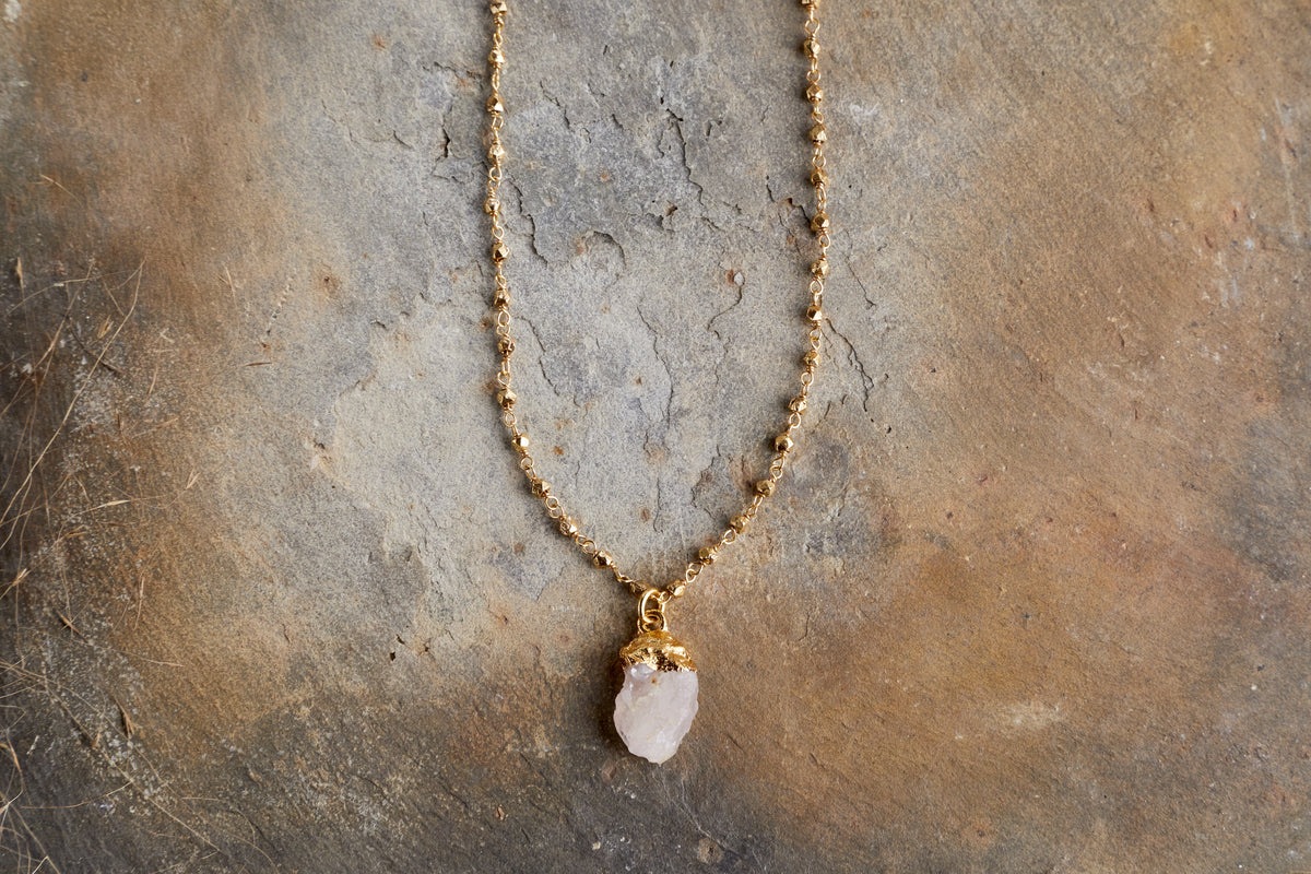 The nkuku Hadari Rose Quartz Necklace—featuring a raw, unpolished crystal pendant on a gold chain—rests on an earthy brown surface, showcasing the elegance of this unique jewellery piece.