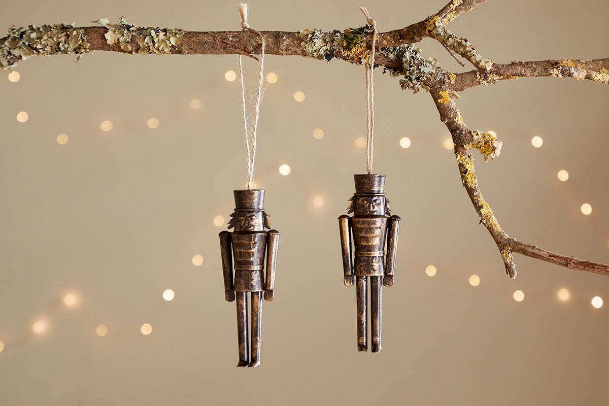 Two Imay Nutcracker Baubles in antique brass by nkuku hang from twine on a mossy branch, with warm white string lights softly glowing in the background—perfect for adding vintage charm to your Christmas tree décor.