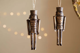 The Imay Nutcracker Baubles - Antique Brass (Set of 2) by nkuku feature simple rectangular soldiers with tall hats, hanging from strings—ideal festive decor for your Christmas tree against a softly lit background.