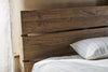 The nkuku Iya Reclaimed Wood Bed - Natural features a rustic wooden headboard with visible grain and cracks, dressed in white sheets and a subtly striped gray pillow, softly lit from the left.