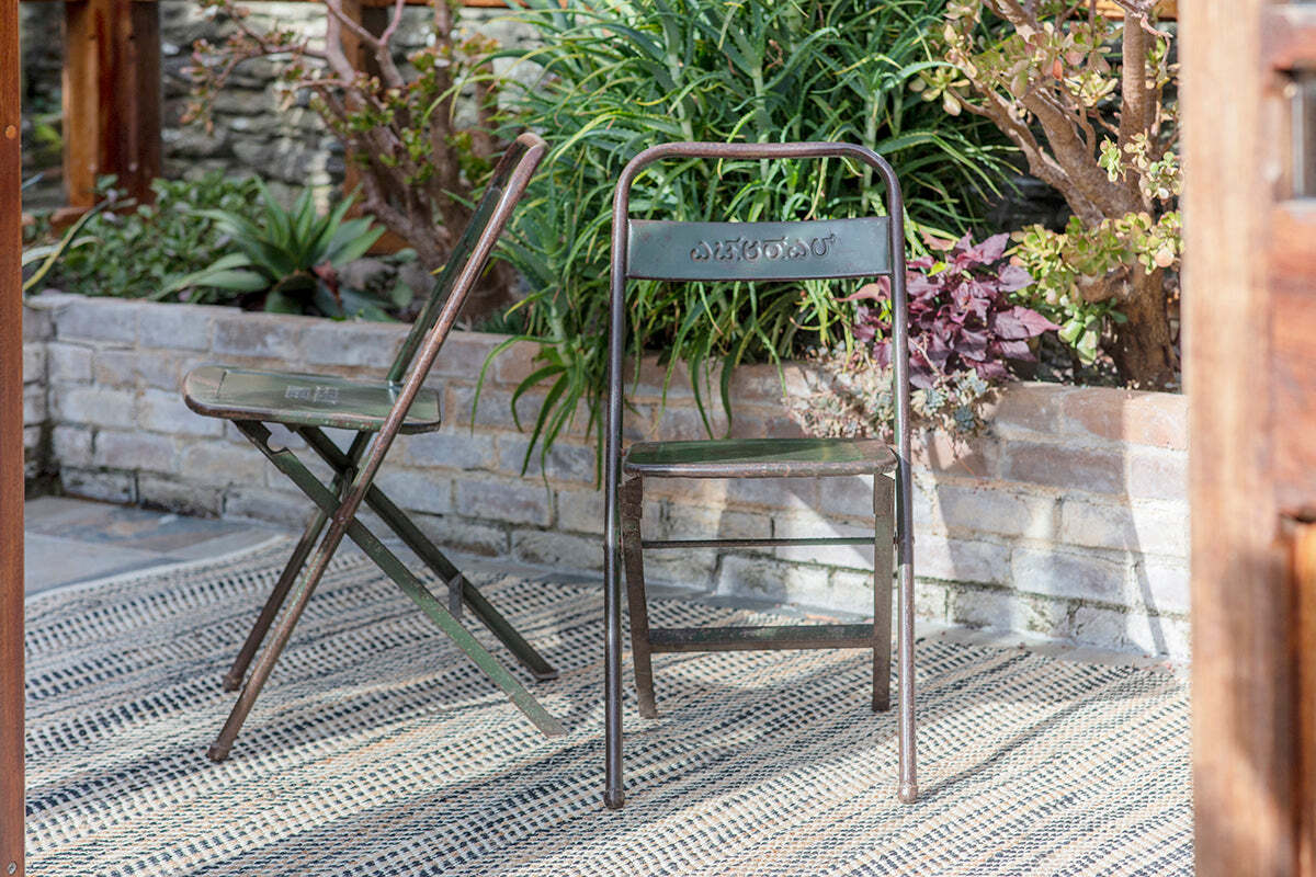 Two nkuku Ishan Reclaimed Folding Chairs—one open, one folded against the wall—rest on a patterned rug. A brick wall and leafy plants complete the rustic look as sunlight gently highlights the garden bed.