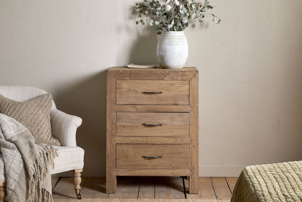 The Iya Reclaimed Wood Chest of Drawers – Natural – Small by nkuku stands beside a beige armchair with a knitted throw. A white vase with green foliage rests atop the dresser, set against a neutral wall and wooden floor.