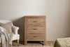 The nkuku Iya Reclaimed Wood Chest of Drawers - Natural - Small, featuring three drawers with metal handles, stands on a wooden floor next to a white armchair draped with a beige knitted throw. A textured beige bedspread is visible on the right.