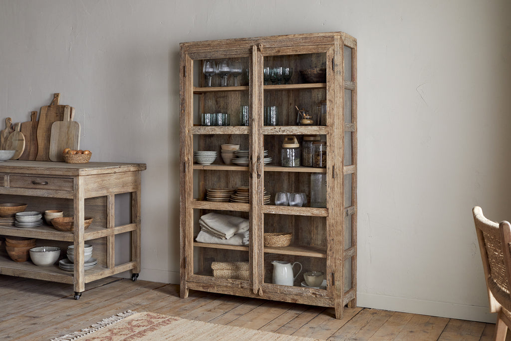 The nkuku Ibo Reclaimed Wood & Glass Cabinet - Natural - Wide displays dishes, bowls, and glassware behind glass panels with antique brass handles. Nearby, a matching side table holds bowls and cutting boards in a cozy, neutral minimalist setting.