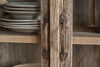 Close-up of the nkuku Ibo Reclaimed Wood & Glass Cabinet - Natural - Wide, showing antique brass handles, metal hardware, and stacked ceramic plates and bowls on its wooden shelves.