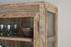 Close-up of the nkuku Ibo Reclaimed Wood & Glass Cabinet - Natural - Wide, featuring glass panels, antique brass handles, and a weathered wood finish. Inside, a wooden bowl and four clear glass goblets are displayed on the shelf.